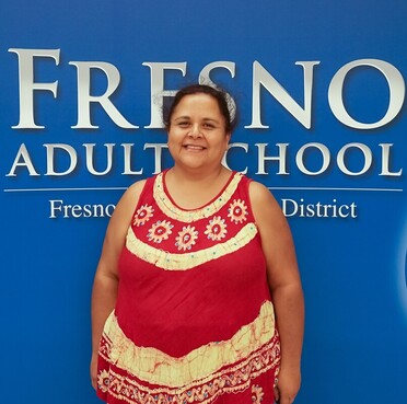 A picture of a smiling female named Ana Maria Bustamante, who is wearing a red and white dress and standing in front of a blue Fresno Adult School sign